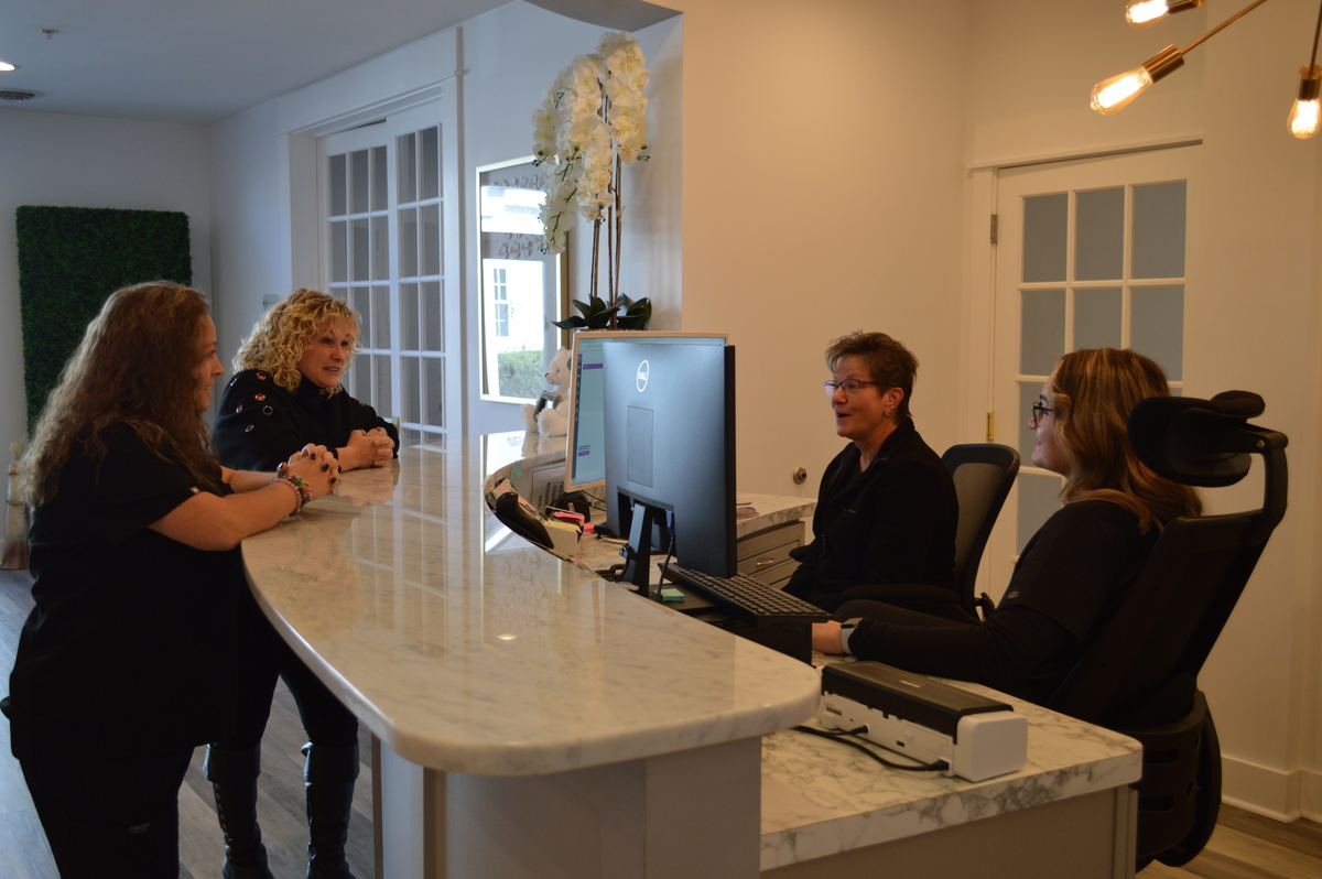 two women standing at the front desk at the fertility wellness institute, talking to the two women sitting behind the desk